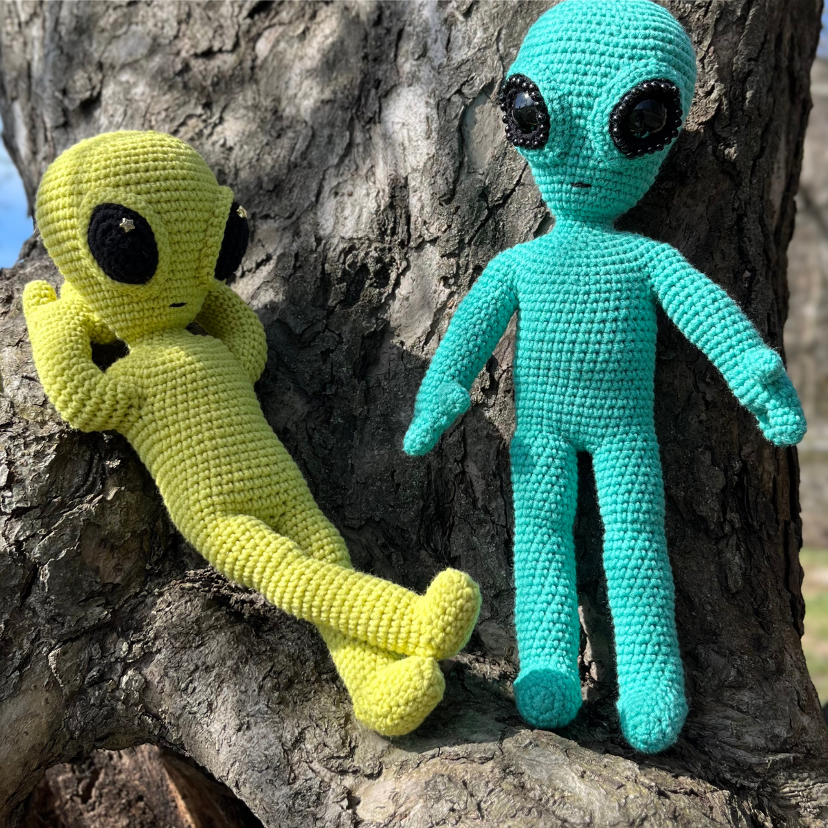 Two crocheted amigurumi alien toys sitting on a tree branch, one in yellow and the other in turquoise with black eyes.