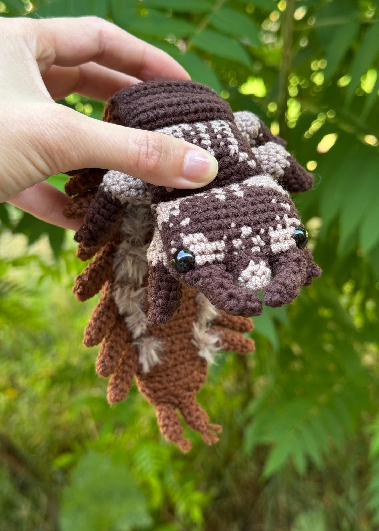A hand holds a crochete hellgrammite a brown and cream patterned against a blurred green outdoor background.