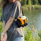 A woman wearing a gray t-shirt and jeans stands outdoors by a lake, carrying a crocheted bumblebee shoulder bag.