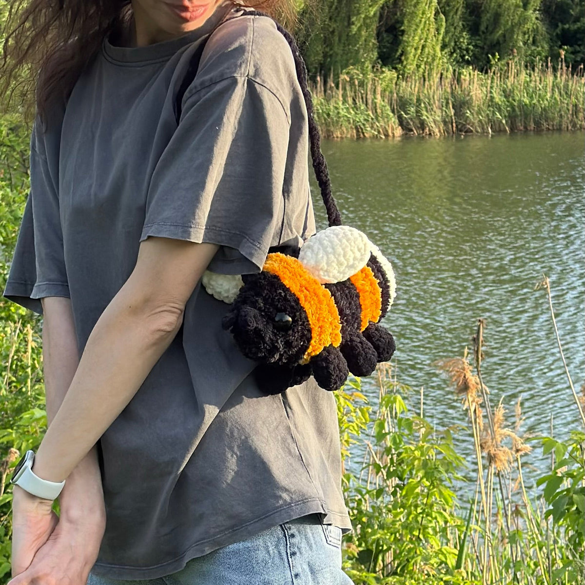 A woman wearing a gray t-shirt and jeans stands outdoors by a lake, carrying a crocheted bumblebee shoulder bag.