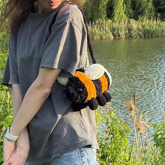 A woman wearing a gray t-shirt and jeans stands outdoors by a lake, carrying a crocheted bumblebee shoulder bag.