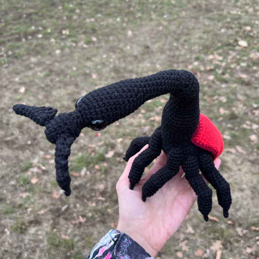A hand holds a black and red crocheted Giraffe Weevil toy with a red shell against an outdoor grassy background.