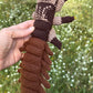 A hand holds a crocheted hellgrammite toy outdoors against a blurred green grassy background with small white flowers. The toy is made of brown, dark brown, and light beige yarn with black bead eyes.