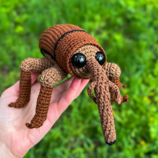A hand holds a crocheted weevil beetle toy outdoors against a blurred green grass background. The toy has a brown body, tan legs, large black eyes, and a long brown nose.