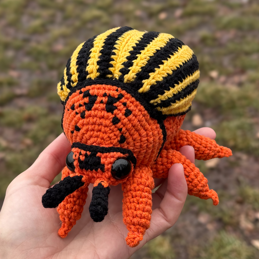 Hand holding a crocheted Colorado beetle with black and yellow stripes against a natural background.